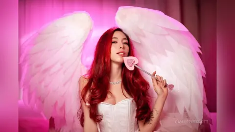 Red-haired angel in a white corset holding a pink heart wand against a dreamy pink background.