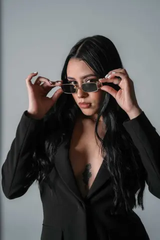 Stylish woman with long dark hair wearing a black blazer and rectangular sunglasses in a minimalist studio setting.