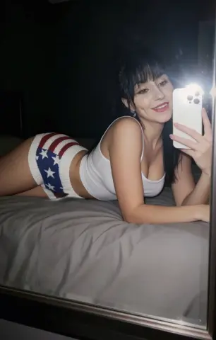 Young woman in white tank top and American flag print shorts taking a selfie on a bed in a moody bedroom.