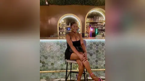 Valjohn sits on a barstool wearing a short black dress, smiling in a bar setting with a tiled counter behind her.