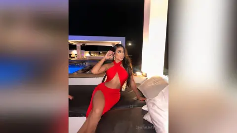 CristalMatthews poses in a red cut-out dress while sitting on a lounge bed by a pool at night.