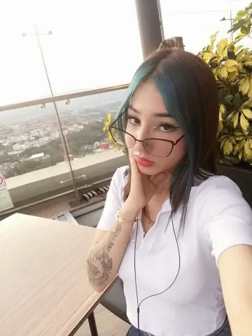 Young woman with blue hair and glasses in a white polo shirt sitting at an outdoor cafe balcony.