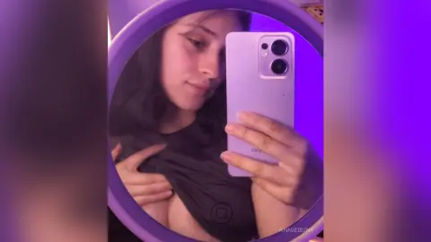 Young woman in a black top taking a moody selfie in a circular mirror with purple lighting.