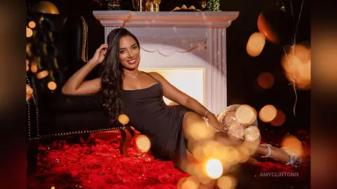 Smiling woman in a black cocktail dress sitting on a red shag rug near a fireplace during a festive party.