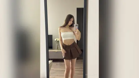 EmorySwift posing in a mirror wearing a white tube top and a short brown skirt, showing her long dark hair and slim build.
