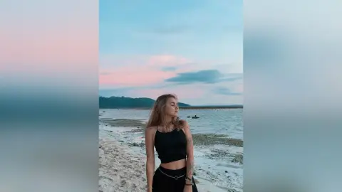 Young woman in a black tank top relaxing on a serene beach at sunset with ocean waves in the background.