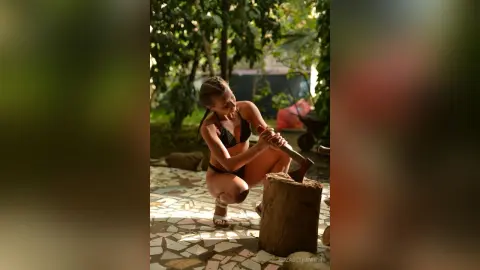 Fit woman in black bikini squatting by a wooden stump in a sunny garden setting.