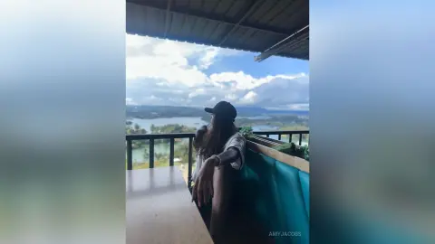 Young woman in a white shirt and black cap relaxing on a balcony overlooking a scenic lake and mountains.