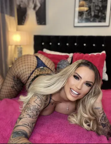 Summer_FoxxX smiles while posing on a pink bed in fishnet stockings and blue lingerie, showing off her blonde hair and tattoos.