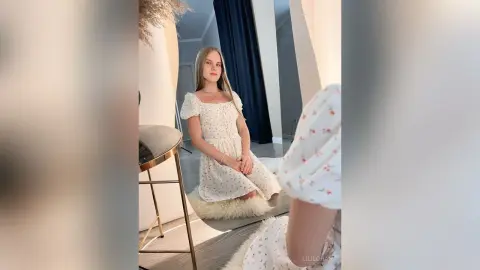 LiliLohan sits on a white furry rug wearing a white floral print sundress with short sleeves.