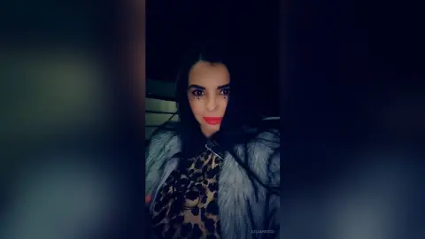 Young woman with long dark hair wearing a leopard print top and grey faux fur coat in a moody nighttime setting.