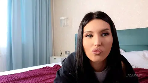 JadaJosey with long dark hair makes a pouty face while sitting on a bed in a bedroom.