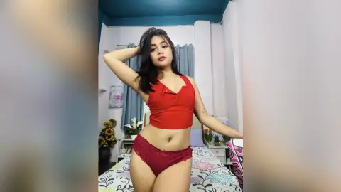 Young woman in red crop top and matching briefs posing in a bright bedroom setting.
