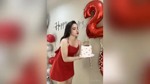 Young woman in a red dress holding a birthday cake next to large red number 2 balloons in a festive setting.