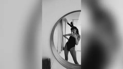Moody black and white selfie of a woman in a black bodysuit and fishnets reflected in a round mirror.