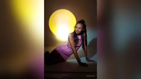 Young woman in a pink sleeveless dress posing against a glowing yellow orb in a moody studio setting.