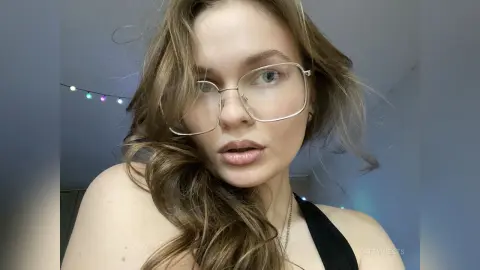 Young woman with blonde hair and gold glasses wearing a black top in a soft, natural indoor setting.