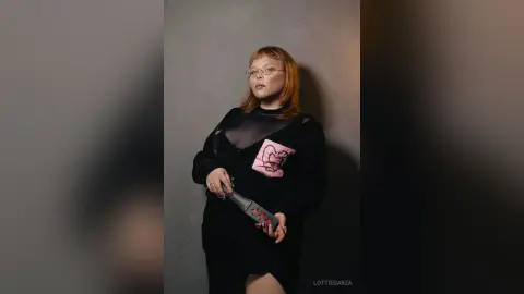 Redheaded woman in a black sheer mesh top and pink graphic tee posing against a neutral studio background.