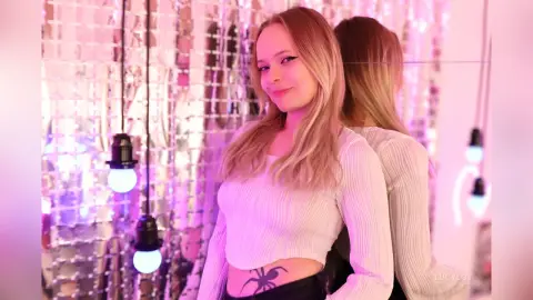 LucyLoy smiles in a white long-sleeve crop top, showing her spider tattoo and blonde hair against a sparkly background.