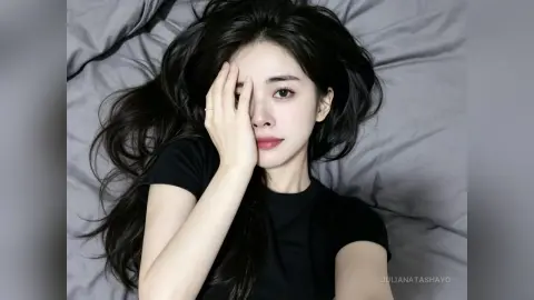 Young woman with long dark hair wearing a black t-shirt lying on grey bedding in a moody, serene setting.