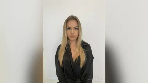Blonde woman in a black satin blouse posing against a minimalist white background with a neutral mood.