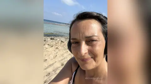 MichaelaBelle smiles while wearing black headphones on a sunny beach.