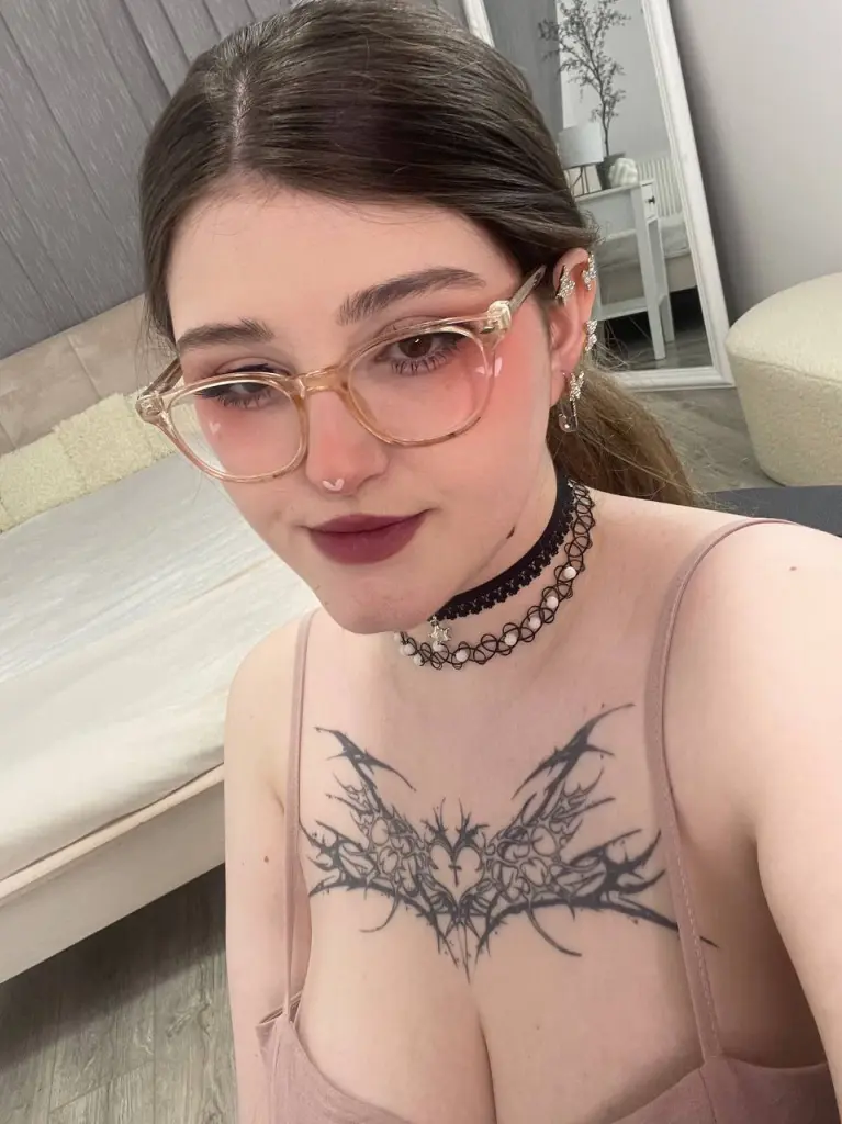 Blonde woman with glasses and a black choker necklace featuring a large chest tattoo in a bright room.
