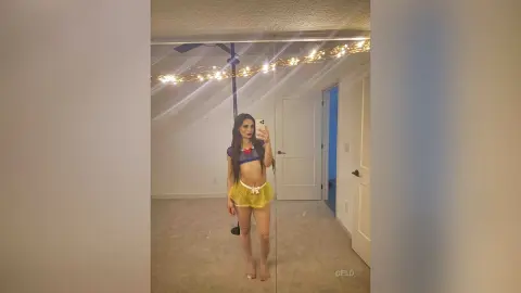 Young woman in a blue crop top and yellow shorts posing for a mirror selfie in a brightly lit bedroom.