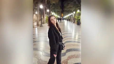 Stylish woman in a black jacket and jeans standing on a patterned stone walkway in a lush tropical park.