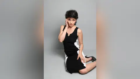 IveteDurand poses sitting on the floor wearing a black and white sleeveless dress with short dark hair against a plain grey background.