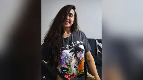 RossiNoleen smiles while wearing a grey graphic t-shirt featuring anime characters, letting her long dark hair fall over her shoulder.