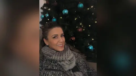 MontseFerrera smiles while wearing a thick grey scarf and a textured grey sweater in front of a lit Christmas tree.