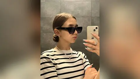 Young woman in a striped shirt and sunglasses taking a stylish mirror selfie in a modern setting.