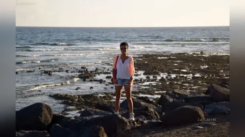 Woman in white top and denim shorts standing on a rocky beach at sunset with ocean waves in the background.