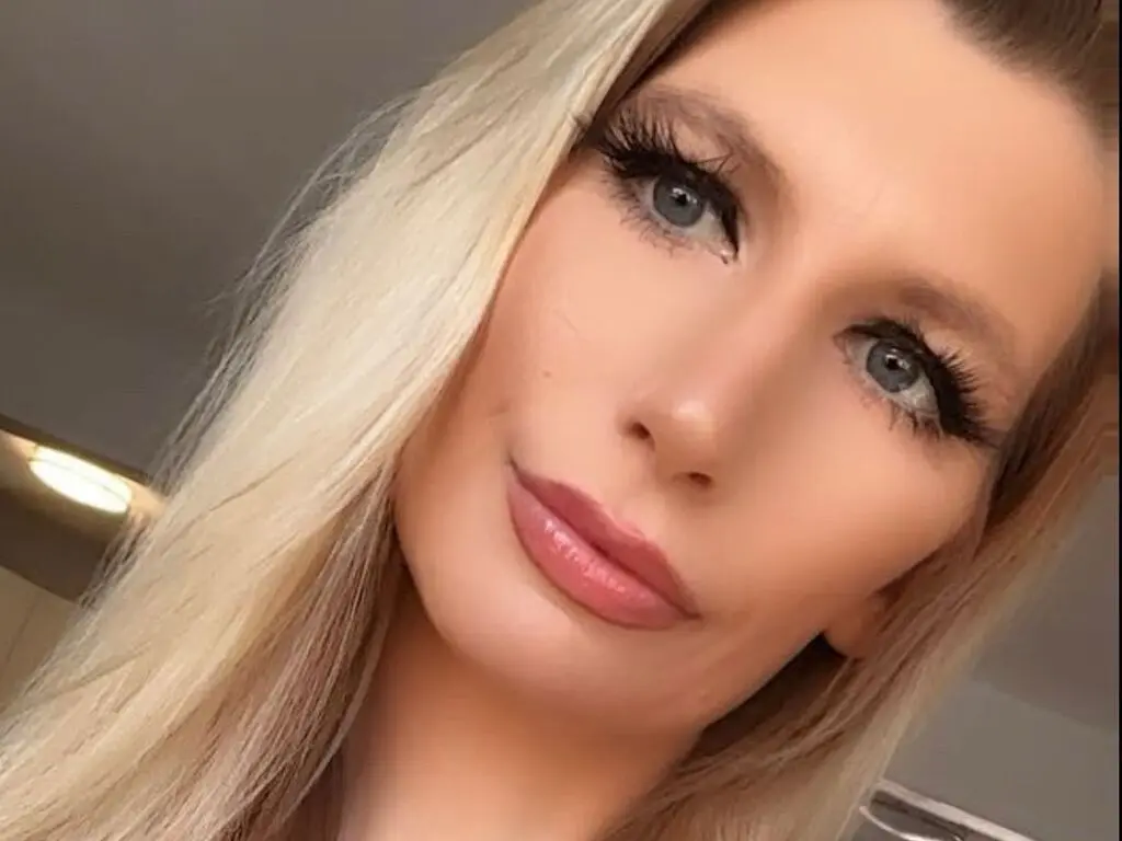 Close-up portrait of a blonde woman with glamorous eye makeup and long lashes looking serene indoors.