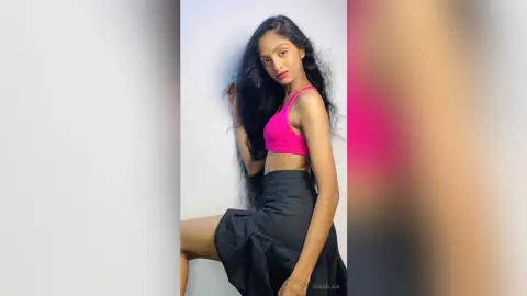 Young woman in a pink crop top and black skirt posing confidently against a white studio background.