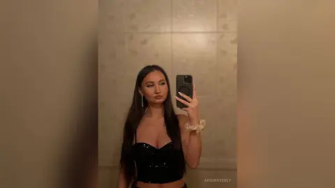 Young woman in a black sequin crop top taking a moody mirror selfie in a modern bathroom.