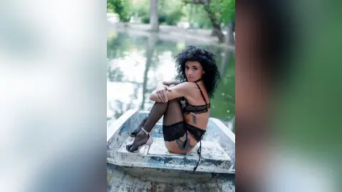 Curvy woman in black lace lingerie and stockings posing on a rustic boat by a serene lake.