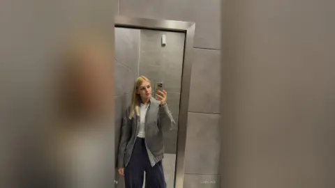 Blonde woman in a grey oversized jacket and navy trousers taking a mirror selfie in a modern grey bathroom.