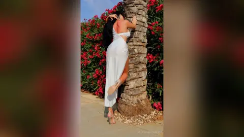Woman in elegant white backless evening gown posing by a palm tree in a lush tropical garden.