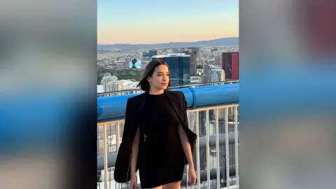 Stylish woman in a black outfit standing on a high-rise balcony overlooking a city skyline at sunset.