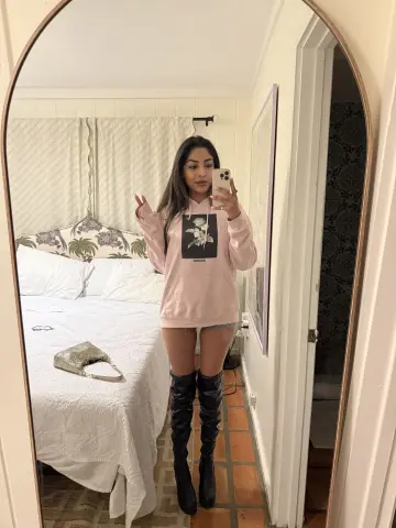 Woman in pink graphic hoodie and black over-the-knee boots taking a mirror selfie in a bright bedroom.