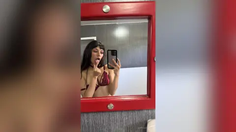 SophiaRichy poses in a red lace bra, sticking her tongue out and holding her phone for a mirror selfie against a grey wall.