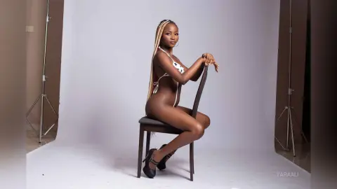 Confident woman with long blonde braids in a white bikini sitting on a chair in a bright photo studio.