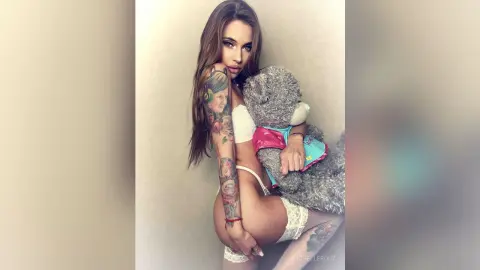 Tattooed woman in white lingerie hugging a large grey teddy bear in a minimalist indoor setting.