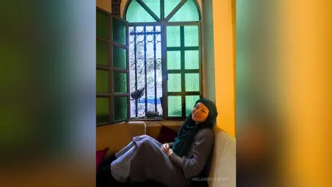 Smiling woman in a black hijab and grey outfit relaxing on a sofa by a sunlit window.