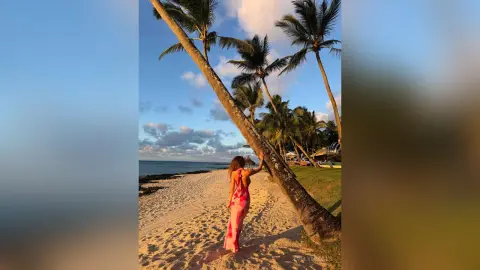 Woman in a pink summer dress walking on a tropical beach under palm trees at sunset.