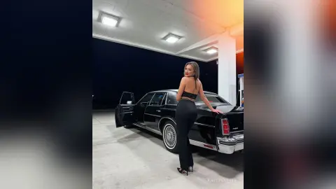 Stylish woman in black jumpsuit posing by a vintage black car in a moody parking garage at night.