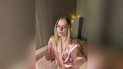 Blonde woman in a pink satin robe relaxing in a cozy, dimly lit bedroom setting.