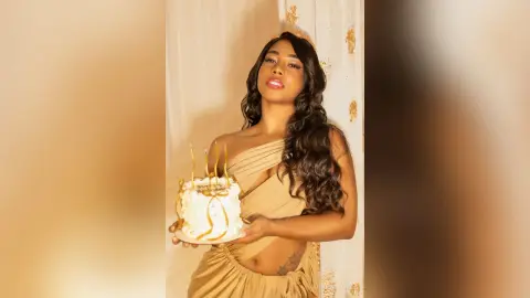 Glamorous woman in a gold satin dress holding a white frosted cake in a warm, elegant setting.